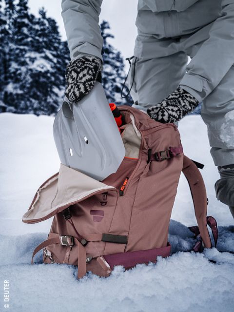 Une personne en vêtements d'hiver et gants à motifs place une pelle à neige dans un sac à dos rose sur un sol enneigé, avec des arbres enneigés en arrière-plan.