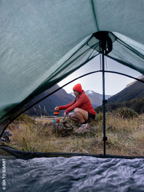 Une personne en veste rouge et bonnet prépare de la nourriture à l'extérieur, accroupie dans l'herbe, près de matériel de camping, vue à travers la porte ouverte d'une tente avec des montagnes et des arbres en arrière-plan.