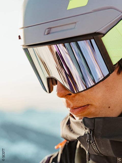 Gros plan sur une personne portant un casque de ski gris, des lunettes de ski réfléchissantes et une veste foncée, avec un paysage enneigé flou en arrière-plan.