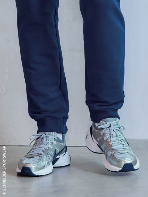 Une personne en jogging bleu marine et chaussures de course grises à semelles blanches se tient à l'intérieur sur un sol clair.