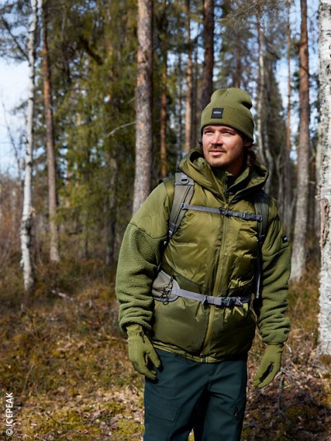 Un homme avec un bonnet, une veste et des gants vert olive se tient dans une forêt ensoleillée et regarde sur le côté. Il porte un sac à dos et est entouré de grands arbres et de maigres broussailles.
