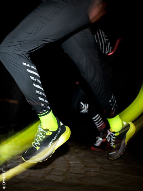 Un gros plan de deux personnes courant la nuit. L'accent est mis sur leurs jambes, vêtues de pantalons de sport noirs, de chaussettes jaune néon et de chaussures de course DYNAFIT jaunes et noires, le flou de mouvement soulignant la vitesse.