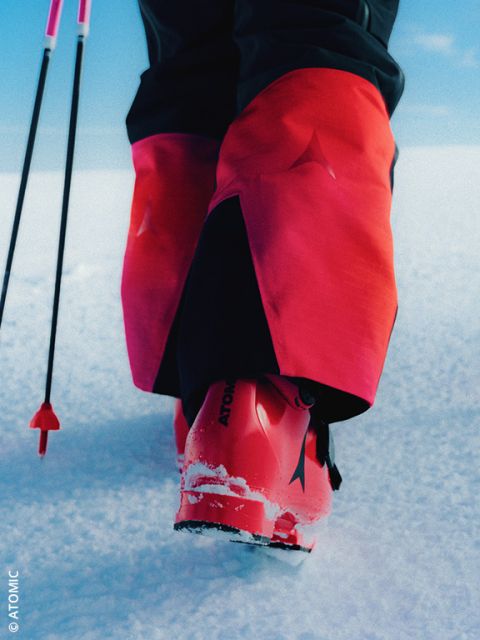 Une personne en pantalon de ski beige clair et en chaussures de ski grises se tient dans la neige avec des bâtons de ski. Les chaussures de ski ont des accents rouges et un logo avec l'inscription "VAN DEER".