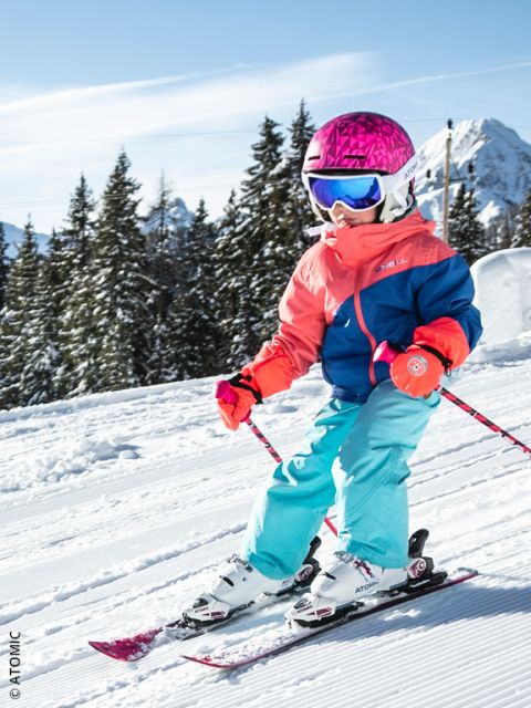 Un jeune enfant portant un casque rose vif, des lunettes bleues et un équipement de ski coloré dévale une piste enneigée, entouré de pins et de montagnes sous un ciel bleu clair.