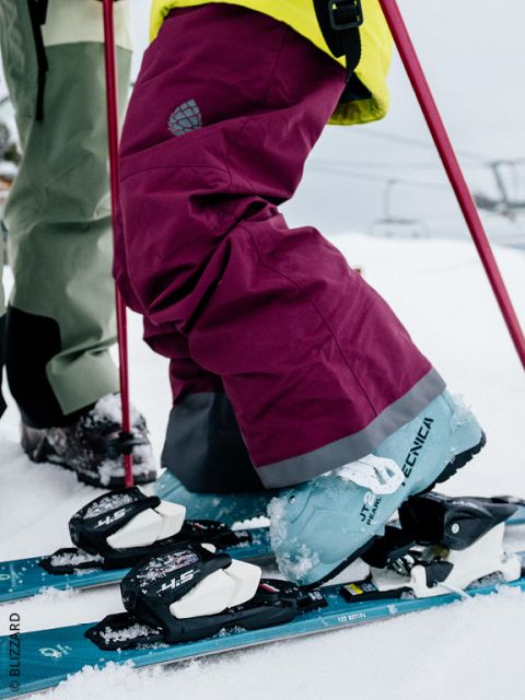 Gros plan sur les jambes d'un skieur portant un pantalon de ski marron et des chaussures de ski Tecnica bleues, clipsées dans les fixations dans la neige, avec des bâtons de ski rouges et un téléski en arrière-plan.