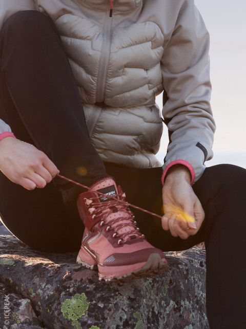Une personne vêtue d'une veste bouffante claire et d'un pantalon noir est assise sur un rocher et lace les lacets d'une chaussure de randonnée marron, tandis que le soleil du soir illumine doucement la scène.