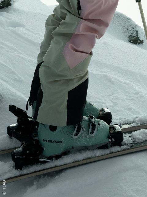 Une personne portant des chaussures de ski HEAD vert menthe et un pantalon de ski beige et rose se tient sur des skis dans la neige fraîche.