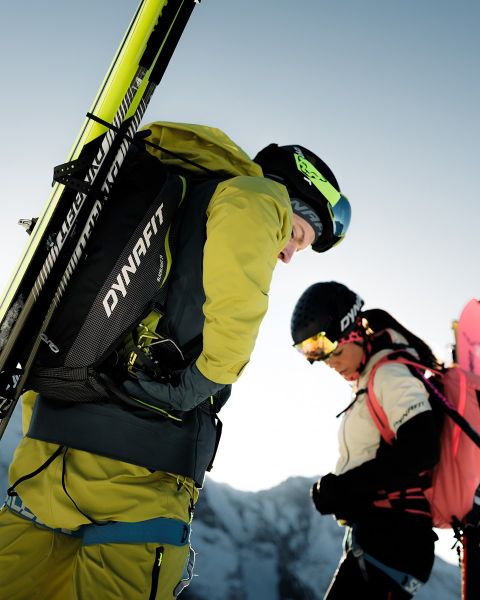 Deux personnes courent sur un sentier de montagne avec une ville et une rivière en arrière-plan. L'une porte un t-shirt noir et un short, l'autre un débardeur bleu et un short. Le logo Dynafit se trouve en haut de l'image.