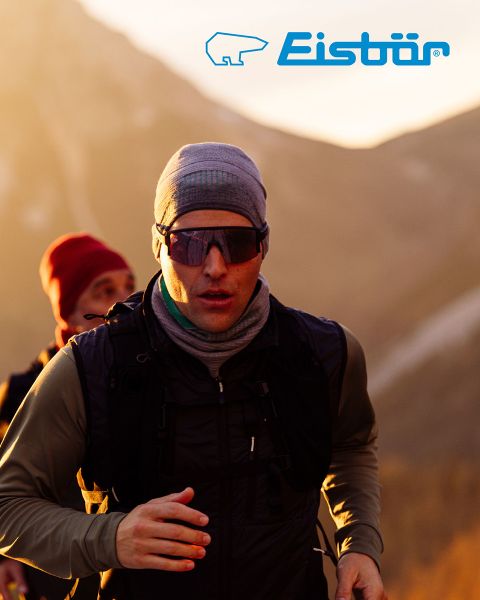 Une femme et un homme sourient en faisant du ski dans un paysage enneigé. Elle porte une veste rouge et un bonnet blanc et tient des bâtons de ski, lui une veste beige et un bonnet vert. Au-dessus d'eux, on voit le logo "Ours polaire".