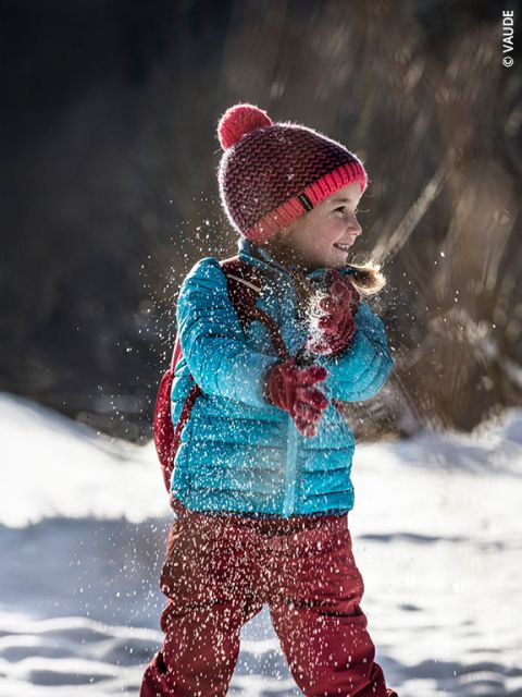 Un jeune enfant en vêtements d'hiver lance de la neige, sourit et joue dehors par une journée ensoleillée et enneigée. L'enfant porte une veste bleue, un pantalon rouge et un bonnet rouge en tricot avec un pompon.