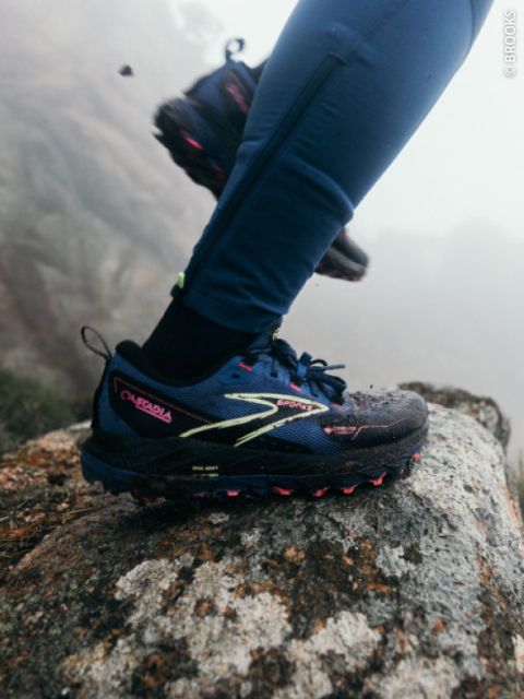Une personne portant des chaussures de trail bleues et des leggings bleus entre dans une zone rocheuse à l'extérieur, avec du brouillard et des rochers en arrière-plan.