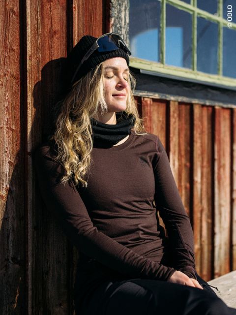 Une personne aux longs cheveux blonds, portant un bonnet noir, des lunettes de soleil et des vêtements sombres, est assise les yeux fermés contre un mur en bois au soleil, semblant détendue et satisfaite.