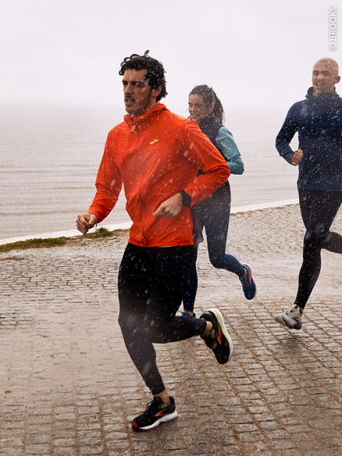 Trois personnes font du jogging par temps pluvieux sur un chemin pavé humide au bord de l'eau. La personne de devant porte une veste rouge, tandis que les deux personnes derrière portent des vêtements plus sombres. La pluie tombe visiblement autour d'eux.