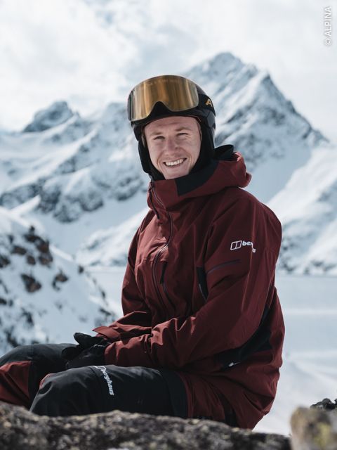 Une personne portant une veste de ski marron, un pantalon de ski noir et un casque avec des lunettes de ski est assise sur une montagne enneigée avec des sommets enneigés en arrière-plan et sourit à la caméra.