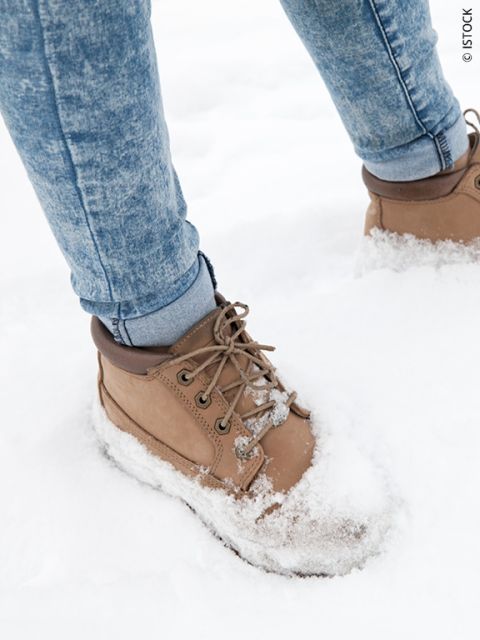 Une personne portant des bottes marron clair et un jean bleu se tient dans la neige fraîche et blanche, la neige recouvrant les bottes et une partie du jean.