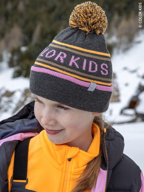 Un enfant souriant avec un bonnet gris sur lequel est écrit « COLOR KIDS » porte une veste jaune et un manteau violet et se tient dehors dans un paysage montagneux enneigé.