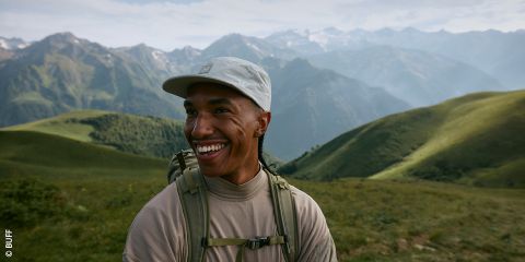 Une personne souriante, portant un bonnet léger et un sac à dos, se tient à l'extérieur dans un paysage de montagne verdoyant avec des sommets lointains et brumeux sous un ciel partiellement nuageux.