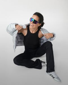 Une femme est assise par terre et enfile un sweat-shirt gris par-dessus sa tête en souriant. Elle porte un haut noir sans manches, un pantalon de jogging gris, des chaussettes blanches et des baskets grises devant un fond gris clair uni.