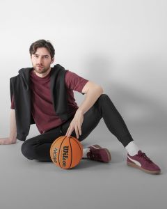 Un homme aux cheveux bruns et à la barbe est assis par terre devant un bloc blanc. Il porte une veste gris clair, une chemise noire, un pantalon de jogging foncé, des chaussettes blanches et des baskets grises, et tient une bouteille d'eau à la main.