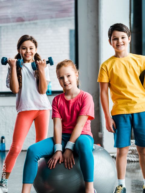 Trois enfants souriants en tenue de sport posent dans un gymnase. Une fille tient des haltères, une autre est assise sur un ballon de gymnastique, et un garçon se tient à côté. Ils ont l'air heureux et prêts pour l'activité physique.