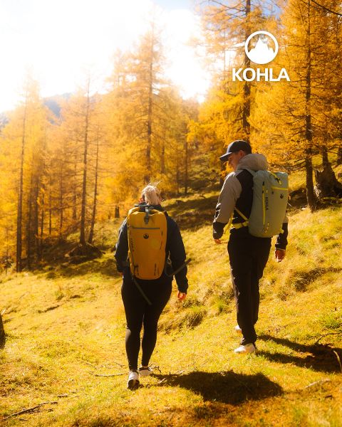 Deux randonneurs avec des sacs à dos marchent à travers une forêt ensoleillée avec des arbres d'automne dorés. La lumière du soleil filtre à travers les branches et projette une lumière chaude. Le logo Kohla est visible dans le coin supérieur droit.