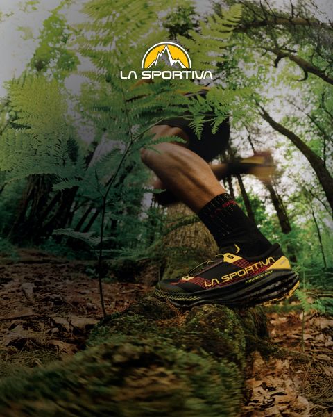 Quatre personnes en équipement de plein air, avec des tapis de bloc sur le dos, montent un sentier de montagne herbeux. Au-dessus d'eux, le logo de La Sportiva est visible dans le ciel. À l'arrière-plan, on aperçoit des sommets montagneux escarpés.
