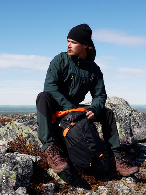 Un homme avec une veste sombre, un bonnet noir et des chaussures de randonnée est assis sur un terrain rocheux à l'extérieur et porte un sac à dos noir et orange. Le ciel est clair et bleu en arrière-plan.