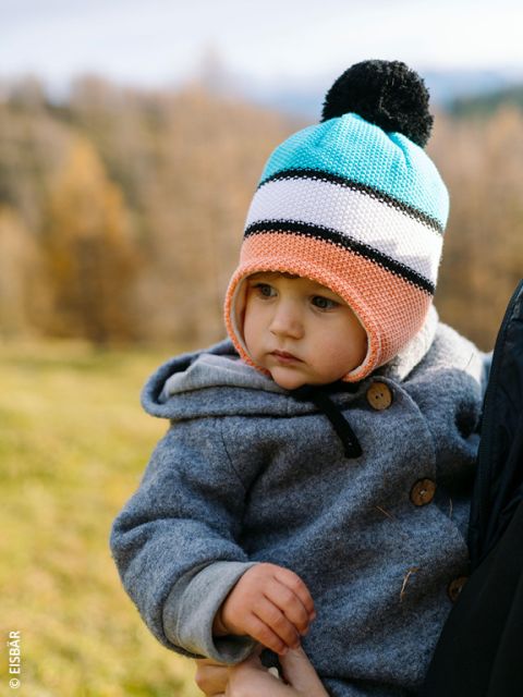 Un jeune enfant portant un bonnet tricoté coloré et un manteau gris est tenu par un adulte à l'extérieur, avec un arrière-plan flou d'arbres d'automne et d'herbe.