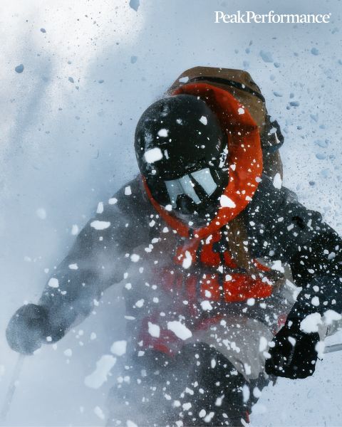 Une personne saute entre de grands rochers dans une région montagneuse sous un ciel bleu éclatant. Elle porte des vêtements de sport, notamment une veste et un short. En haut, on peut lire l'inscription « Peak Performance ».
