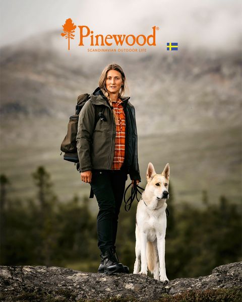 Un homme et une femme en vêtements de plein air marchent dans une prairie sous un ciel nuageux. La femme porte un sac noir, tandis que l'homme tient un chapeau. Au-dessus d'eux, on peut voir le logo Pinewood et le texte « Scandinavian Outdoor Life ».