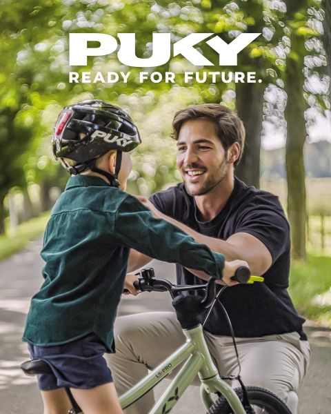 Une jeune fille avec un casque bleu et un t-shirt rayé fait du vélo bleu clair par une journée ensoleillée. Souriante, elle passe devant des graffitis colorés dans un environnement urbain. Le logo PUKY et le slogan « Ready for life » sont visibles en haut à gauche.