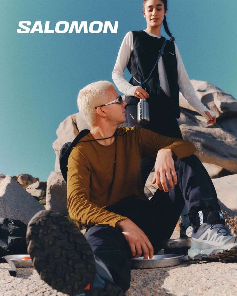 Gros plan d'une personne portant des chaussures Salomon et des chaussettes Salomon avec un dégradé de couleurs bleues. Elle se tient dans une prairie avec un paysage de montagne pittoresque en arrière-plan. Un randonneur en vêtements d'extérieur se tient plus loin, également dans la prairie.