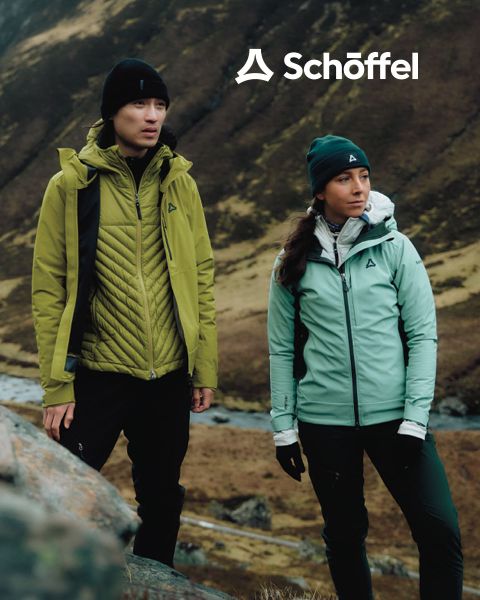 Deux personnes en équipement de plein air marchent sur une colline herbeuse avec des montagnes et des nuages en arrière-plan. Au-dessus d'eux se trouve le logo Schöffel, en dessous une ligne ondulée verte décorative.