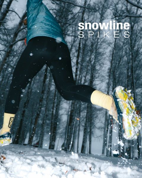 Une personne portant des crampons à neige sur ses chaussures marche dans une forêt enneigée avec de grands arbres sans feuilles. Le texte « snowline SPIKES » apparaît dans le coin supérieur droit. La neige tombe et le sol est recouvert de blanc.