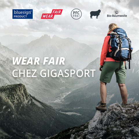 Un randonneur avec un sac à dos se tient sur un promontoire montagneux et contemple une vaste vallée brumeuse. Le texte sur l'image est : "WEAR FAIR CHEZ GIGASPORT" avec des logos pour la durabilité et des étiquettes de marque en haut.