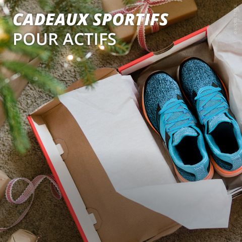 Une boîte de chaussures de sport bleues est ouverte sur le sol à côté d'un sapin de Noël décoré et de cadeaux emballés. Le texte blanc en français se lit comme suit : "Cadeaux sportifs pour actifs".