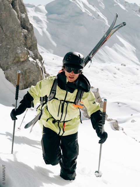 Un skieur avec un casque, des lunettes et une veste jaune gravit une pente de montagne enneigée, portant ses skis sur son dos et utilisant des bâtons de ski pour se soutenir. Des falaises rocheuses et de la neige profonde entourent la scène.