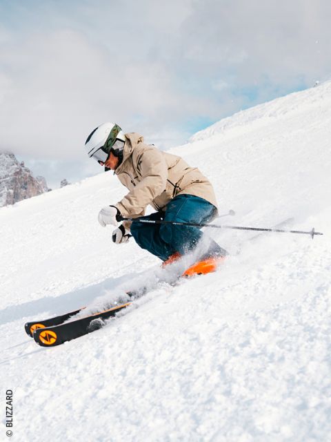 Un skieur en veste beige, pantalon bleu et casque blanc dévale une piste enneigée, effectue un virage serré et soulève de la neige. Des montagnes sont visibles en arrière-plan.