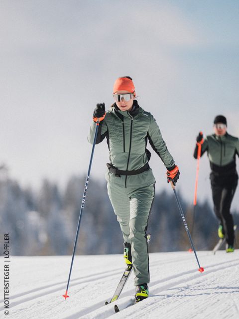 Deux fondeurs sur une piste de ski de fond enneigée, avec équipement de sports d'hiver et lunettes de soleil, avec des arbres et un ciel bleu en arrière-plan.
