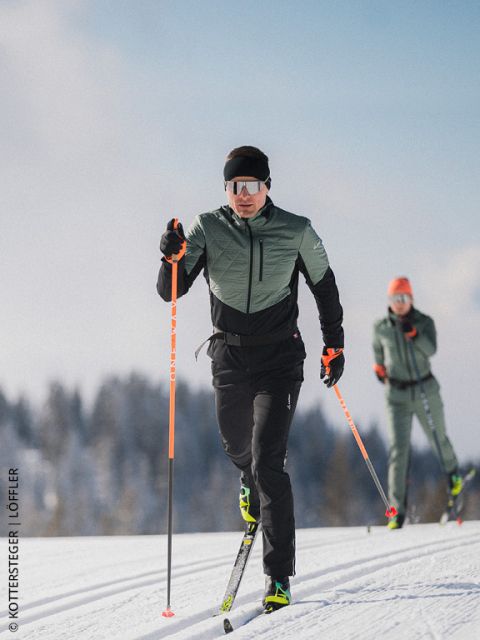 Deux fondeurs sur une piste de ski de fond enneigée, avec équipement de sports d'hiver et lunettes de soleil, avec des arbres et un ciel bleu en arrière-plan.
