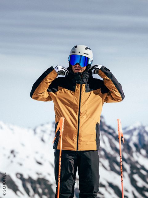 Un skieur en veste jaune et noire ajuste son casque et ses lunettes de ski, debout sur une piste enneigée avec des montagnes en arrière-plan.
