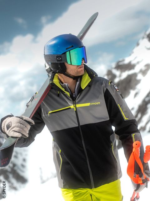 Un skieur avec un casque bleu et des lunettes réfléchissantes se tient sur une montagne enneigée, porte des skis sur l'épaule et tient des bâtons de ski orange.