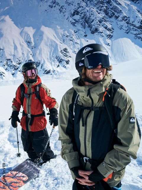 Deux snowboarders en équipement d'hiver se tiennent dans la neige, avec des montagnes enneigées en arrière-plan, sourient et profitent de la journée ensoleillée et lumineuse.