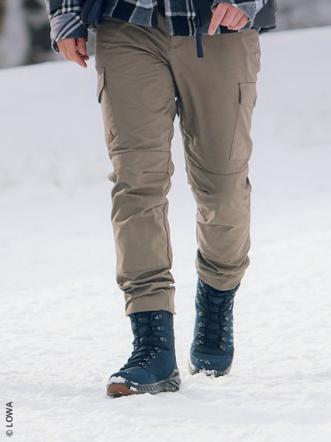 Personne portant un pantalon kaki et des bottes d'hiver bleu marine marchant dans la neige, seuls ses jambes et une partie de son torse étant visibles.