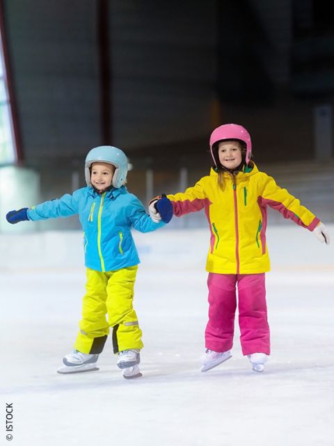 Deux jeunes enfants en vestes et casques colorés patinent ensemble, se tiennent la main et sourient sur une patinoire.
