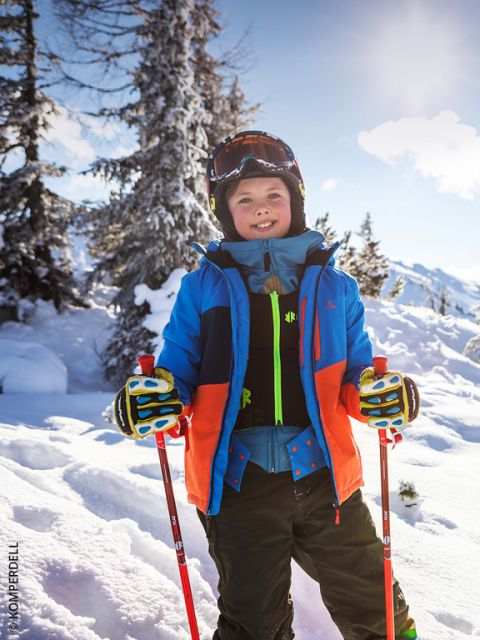 Enfant souriant en équipement de ski avec des bâtons debout dans un paysage de montagne enneigé avec des arbres et une lumière vive du soleil en arrière-plan.