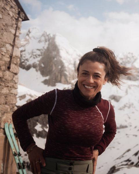 Une femme souriante portant un haut marron châtaigne se tient à l'extérieur près d'un bâtiment en pierre avec des montagnes enneigées et des skis en arrière-plan.