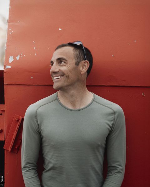 Un homme souriant en chemise verte se tient devant une grande construction métallique rouge et regarde sur le côté avec des lunettes de soleil sur la tête.