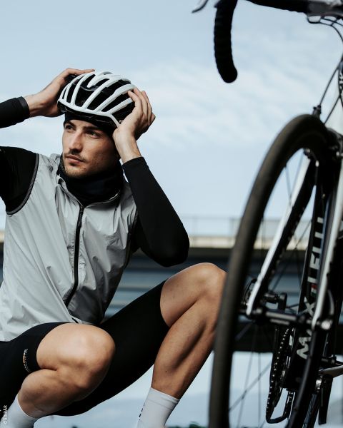 Un cycliste avec un casque et des vêtements de cyclisme s'agenouille à côté d'un vélo et ajuste son casque, avec un pont et le ciel en arrière-plan.