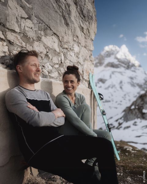 Deux personnes en vêtements d'hiver sont assises à l'arrière d'un véhicule garé sur une route enneigée. Les deux sourient. La personne à gauche porte un bonnet noir et un sweat-shirt, tandis que la personne à droite porte un pull à motifs, un casque et des lunettes de protection. Texte sur l'image.